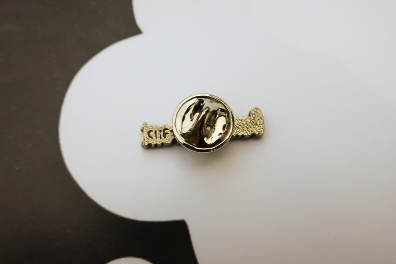 May include: A silver pin with the word "Love" written in a stylized font. The pin has a circular back with a safety clasp.