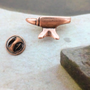 May include: A copper-colored metal anvil pin with a round back. The pin is about 1 inch tall and 1 inch wide.