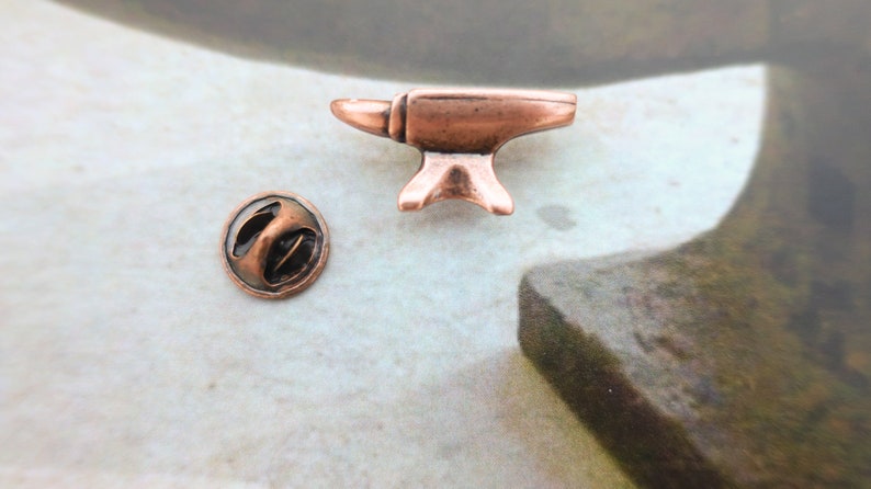 May include: A copper-colored metal anvil pin with a round back. The pin is about 1 inch tall and 1 inch wide.