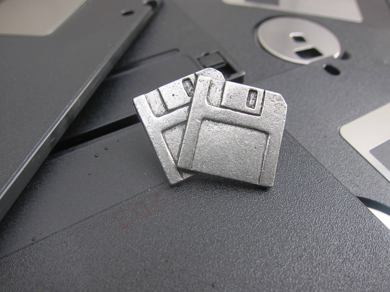 May include: Two silver-colored floppy disk charms are displayed on a dark gray surface, with other floppy disks in the background. The charms are detailed with rectangular and square shapes, and a slot at the top.