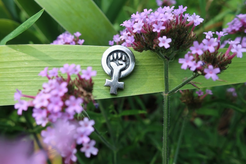 This pewter lapel pin is a raised fist inside the female symbol.  It is placed in a wild flower garden stuck through a tiger lily stalk next to purple wild flowers.