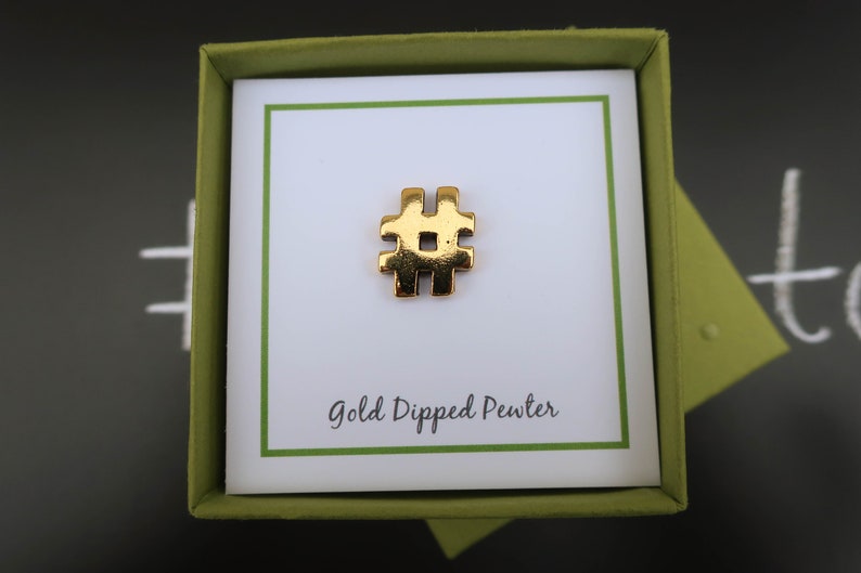 May include: A gold-dipped pewter hashtag charm in a green gift box. The box has a white card inside with the text "Gold Dipped Pewter".
