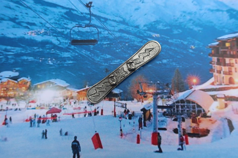 Puede incluir: Un amuleto de snowboard de metal plateado con un dise&ntilde;o de rayo. El amuleto est&aacute; sobre un fondo de una escena de monta&ntilde;a nevada con un remonte.