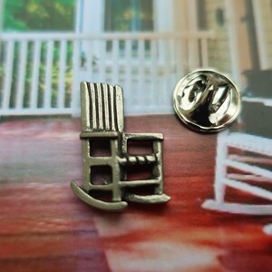 Pode incluir: Um alfinete de cadeira de balan&ccedil;o prateado. O alfinete &eacute; uma r&eacute;plica em miniatura de uma cadeira de balan&ccedil;o com um design detalhado. O alfinete est&aacute; sobre uma superf&iacute;cie vermelha e marrom.