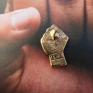 May include: Gold-colored metal pin shaped like a raised fist with the word "CLIFF" inscribed on the back.