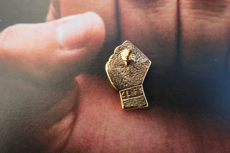 May include: Gold-colored metal pin shaped like a raised fist with the word "CLIFF" inscribed on the back.