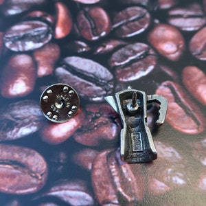 May include: Silver-toned metal pin of a coffee pot with the words "Made in USA" on the bottom. The pin is on a background of coffee beans.