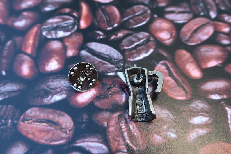 May include: Silver-toned metal pin of a coffee pot with the words "Made in USA" on the bottom. The pin is on a background of coffee beans.
