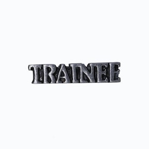 May include: A silver-colored metal pin with the word "TRAINEE" in capital letters. The pin has a slightly distressed, vintage appearance, and is set against a plain white background.