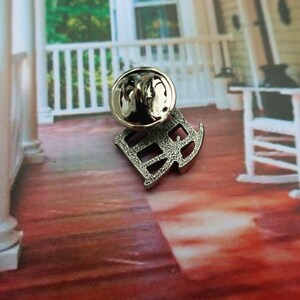 Pode incluir: Pin de metal prateado com uma letra 'R' estilizada com uma superf&iacute;cie texturizada. O pin est&aacute; sobre uma superf&iacute;cie de madeira vermelha.
