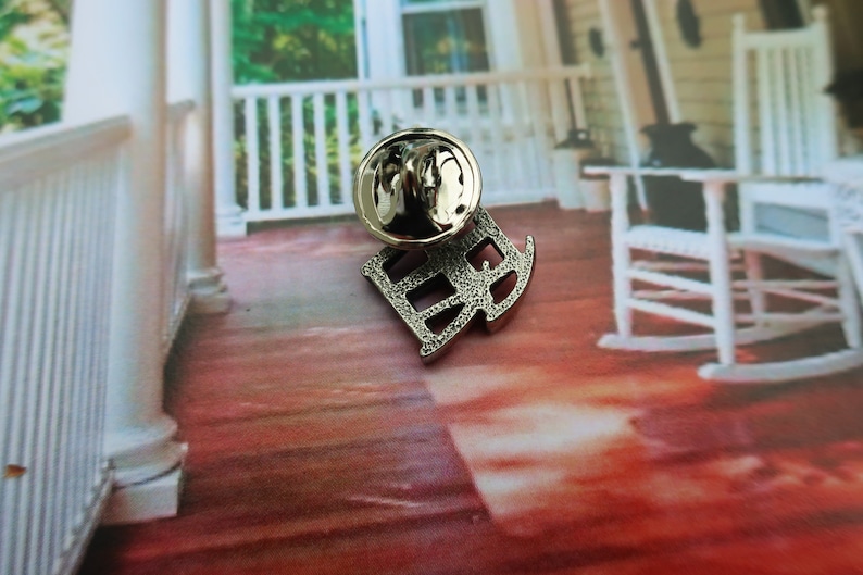Pode incluir: Pin de metal prateado com uma letra 'R' estilizada com uma superf&iacute;cie texturizada. O pin est&aacute; sobre uma superf&iacute;cie de madeira vermelha.