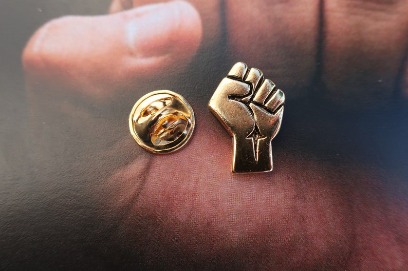 May include: Gold-colored metal pin with a raised fist design. The fist is clenched with the thumb on top. The pin has a simple, circular back.