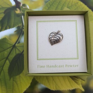 May include: A silver-colored leaf-shaped pewter pin, presented in a green box with a white card. The card reads "Fine Handcast Pewter". The background features blurred green leaves.