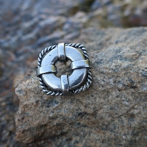 May include: A silver-toned life preserver charm with a rope design. The charm is sitting on a gray rock.