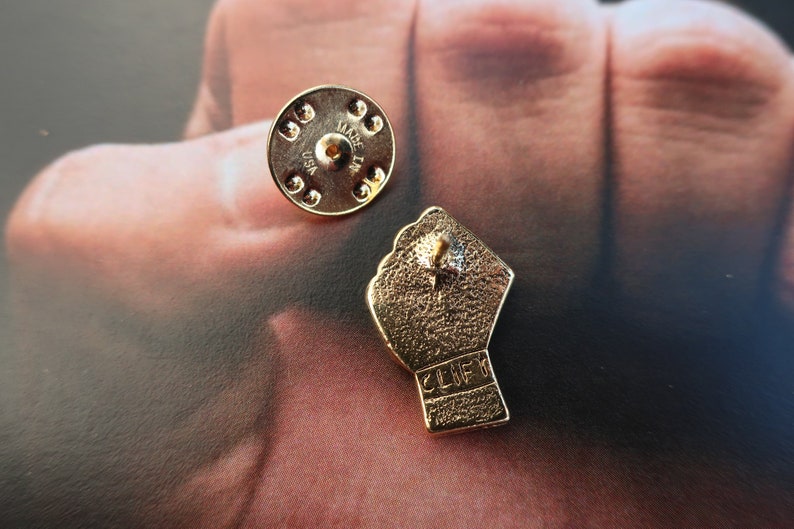 May include: Gold-colored metal pin with a raised fist design. The back of the pin has the text "MADE IN USA" and a pin back.