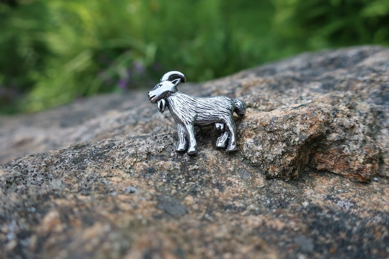 May include: A silver-colored goat figurine, standing on a gray rock. The goat has a detailed, textured surface and is facing to the right.
