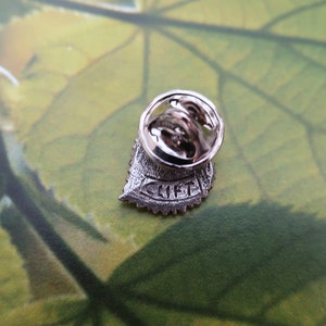 May include: A silver-colored metal pin with a leaf design. The pin has a round back with a clasp and the word "CLIFF" is engraved on the leaf. The pin is set against a green and yellow background.