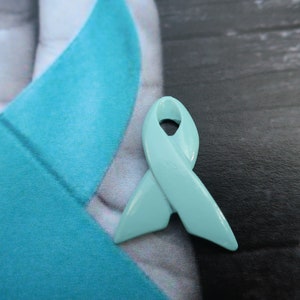 May include: A light blue ribbon, a symbol of awareness and support, is pinned to a turquoise fabric background.