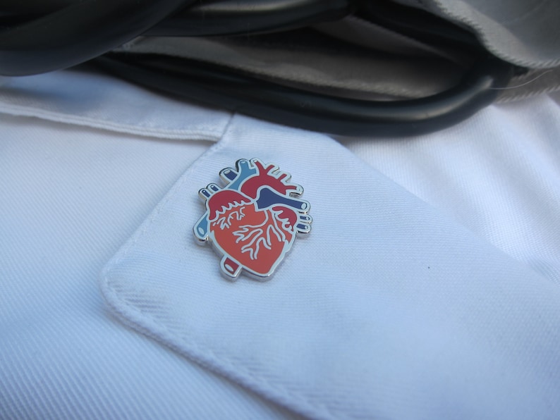 May include: A silver anatomical heart pin with red, blue and white details. The pin is on a white lab coat.