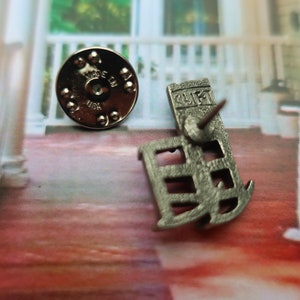 Pode incluir: Pin de metal prateado com um design detalhado de um edif&iacute;cio com a palavra "GULF" na frente. A parte de tr&aacute;s do pin tem um suporte circular de metal com as palavras "MADE IN USA".