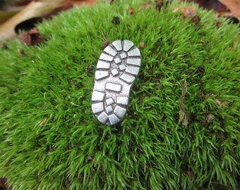 May include: A silver metal charm in the shape of a shoe sole, sitting on a bed of green moss.