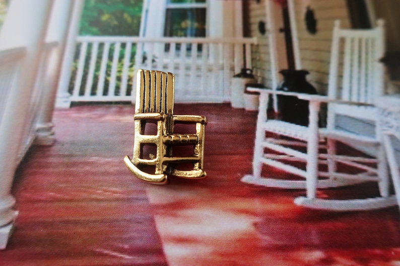 Pode incluir: Um amuleto de cadeira de balan&ccedil;o de metal dourado. O amuleto &eacute; uma r&eacute;plica em miniatura de uma cadeira de balan&ccedil;o tradicional. O amuleto est&aacute; sentado em uma superf&iacute;cie com padr&atilde;o vermelho e branco.