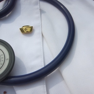May include: A white lab coat with a stethoscope and a gold nurse's cap pin. The stethoscope has a dark blue tube and a silver-colored head with the text "LANN QUALITY TM" on it.