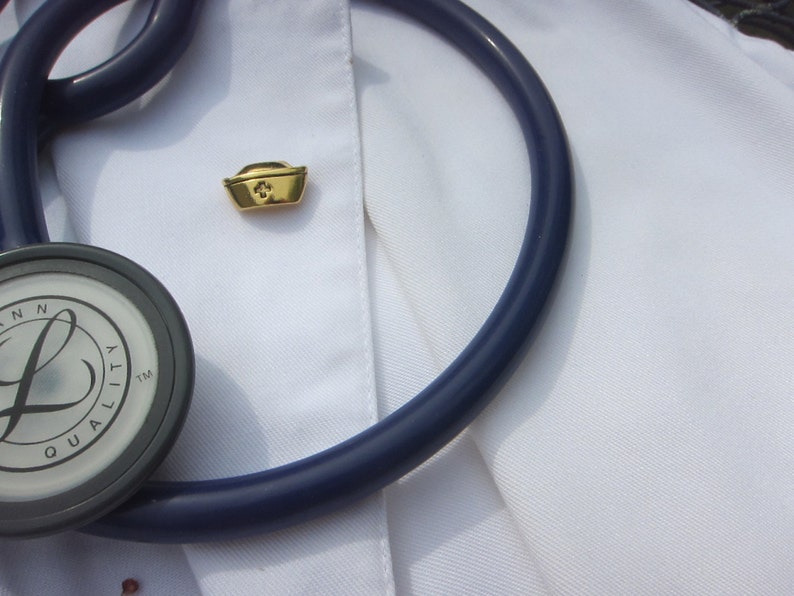 May include: A white lab coat with a stethoscope and a gold nurse's cap pin. The stethoscope has a dark blue tube and a silver-colored head with the text "LANN QUALITY TM" on it.