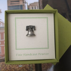 Puede incluir: Un peque&ntilde;o amuleto de plata de la Campana de la Libertad en una caja de regalo verde con el texto "Fine Handcast Pewter".