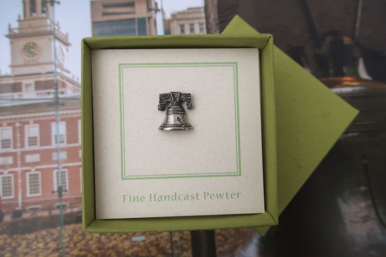 Puede incluir: Un peque&ntilde;o amuleto de plata de la Campana de la Libertad en una caja de regalo verde con el texto "Fine Handcast Pewter".