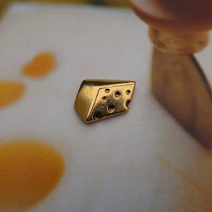 May include: A small, gold-colored, cheese-shaped charm with holes. The charm is on a white surface.