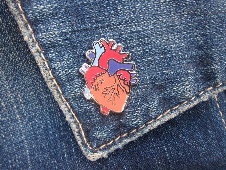 May include: A red, blue and orange anatomical heart enamel pin. The pin is on a blue denim background.