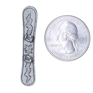 Puede incluir: Un amuleto de metal plateado con forma de tabla de snowboard con un dise&ntilde;o de rayo y tres estrellas. El amuleto es del tama&ntilde;o de una moneda de 25 centavos de d&oacute;lar estadounidense.