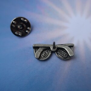 Puede incluir: Pin de gafas de sol de peltre plateado con las palabras "SLIFT" y "PEWTER" en las lentes.