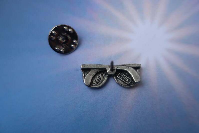 Puede incluir: Pin de gafas de sol de peltre plateado con las palabras "SLIFT" y "PEWTER" en las lentes.