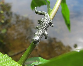 Puede incluir: Un amuleto de cocodrilo plateado colgando de un tallo de planta verde. El cocodrilo está mirando a la cámara con la boca abierta.
