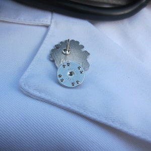 May include: A silver pin featuring an intricate flower design. The reverse side of the pin displays the text "MADE IN ITALY" on a circular metal piece.