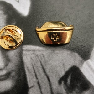 May include: Gold-toned nurse's hat pin with a red cross. The pin has a circular back with a safety clasp.