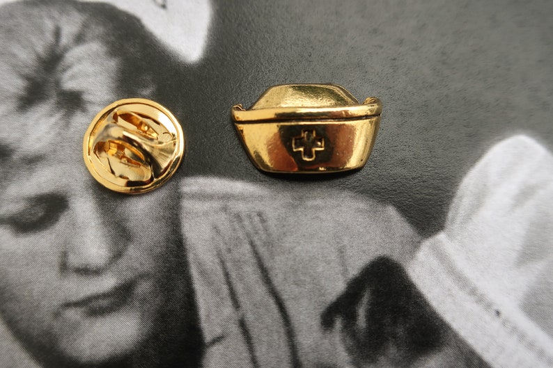 May include: Gold-toned nurse's hat pin with a red cross. The pin has a circular back with a safety clasp.