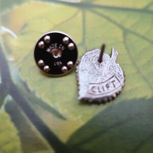 May include: Two metal pins. One is a round black pin back with the text "MADE IN USA". The other is a leaf-shaped pin with the text "CLIFT". The pins are silver in color.