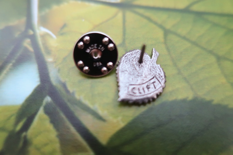 May include: Two metal pins. One is a round black pin back with the text "MADE IN USA". The other is a leaf-shaped pin with the text "CLIFT". The pins are silver in color.
