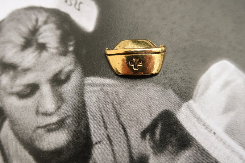 May include: A gold-colored nurse's hat charm with a red cross in the center. The charm is on a white background.