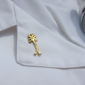 May include: A gold-colored pin shaped like a stylized tree with a flower at the top. The pin is on a white lab coat.
