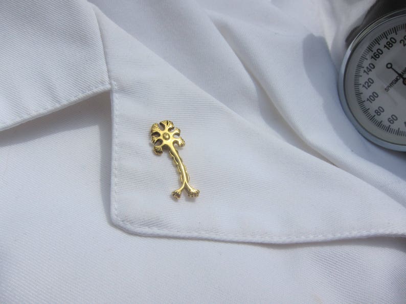 May include: A gold-colored pin shaped like a stylized tree with a flower at the top. The pin is on a white lab coat.