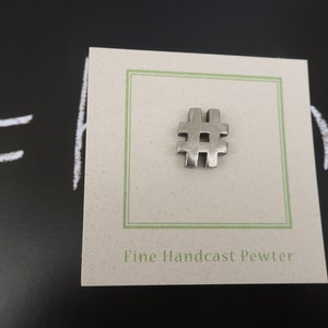 May include: A silver pewter charm in the shape of a hashtag symbol. The charm is displayed on a white card with green trim and the text "Fine Handcast Pewter".
