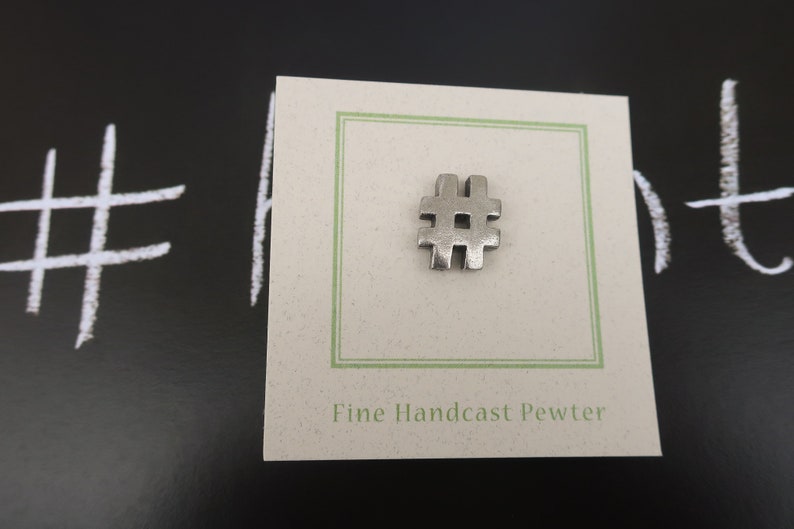 May include: A silver pewter charm in the shape of a hashtag symbol. The charm is displayed on a white card with green trim and the text "Fine Handcast Pewter".