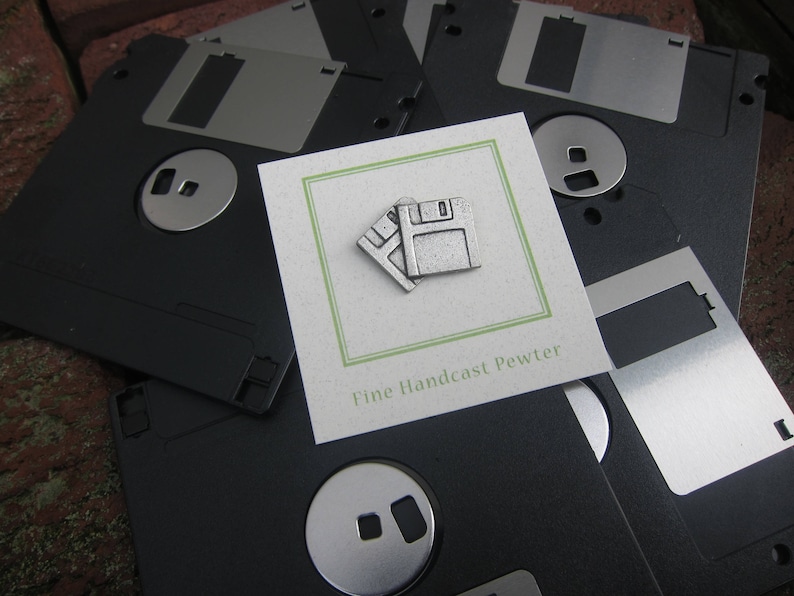May include: A collection of black 3.5-inch floppy disks with metal sliding covers. A small card with a green border displays a pewter floppy disk pin. The card reads "Fine Handcast Pewter".