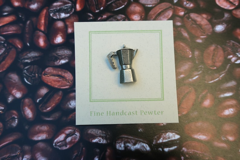 May include: A silver-colored pewter Moka pot pin on a white card with the text "Fine Handcast Pewter". The card is on a background of coffee beans.