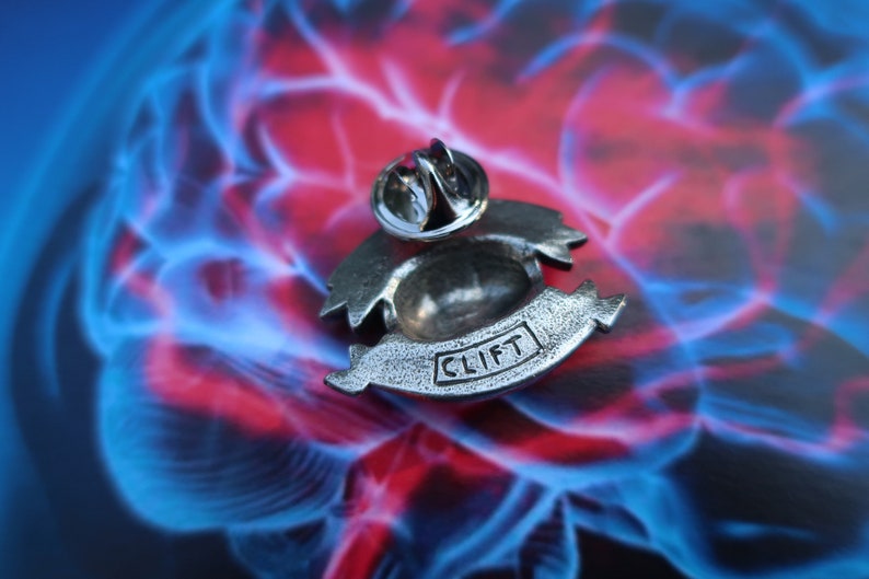 K&ouml;nnte beinhalten: Silberfarbener Anstecker mit einem stilisierten Design, das ein Banner mit dem Wort "CLIFT" zeigt. Der Anstecker befindet sich auf einem blauen und roten Hintergrund.