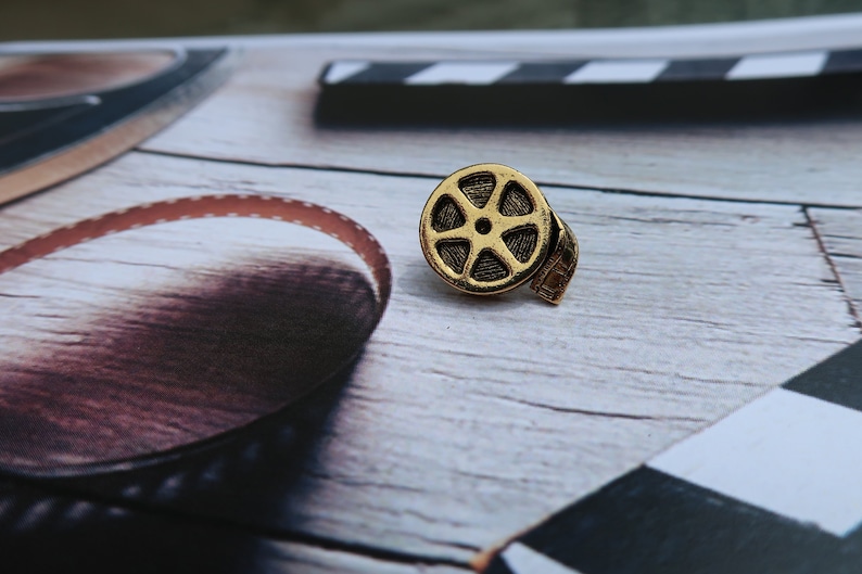 May include: A gold-colored film reel pin with a black and white film strip attached. The pin is on a wooden surface with a black and white checkered pattern in the background.